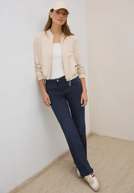Frau mit heller Jacke, weißem Top, blauer Hose und Kappe lehnt an der Wand.